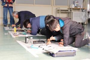 児童とその隣で書き初めをしている地域の方と先生の写真